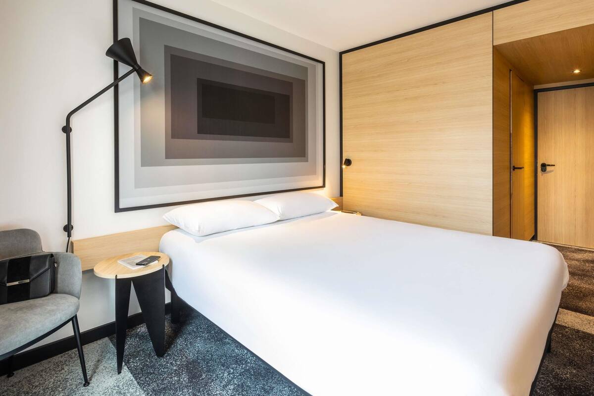 A modern hotel room with a large bed, a desk with a chair, a framed artwork on the wall, and a wardrobe.