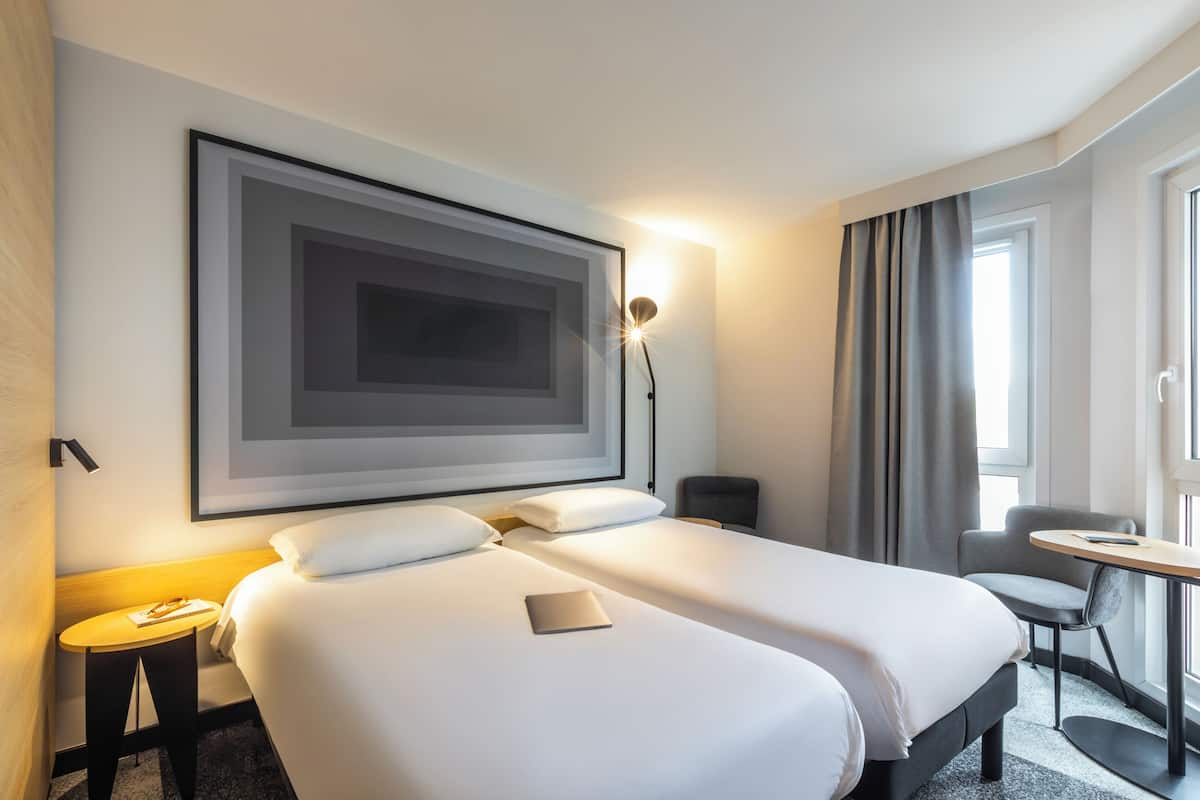 A hotel room with two beds, a desk, a chair, a framed artwork, and a lamp.