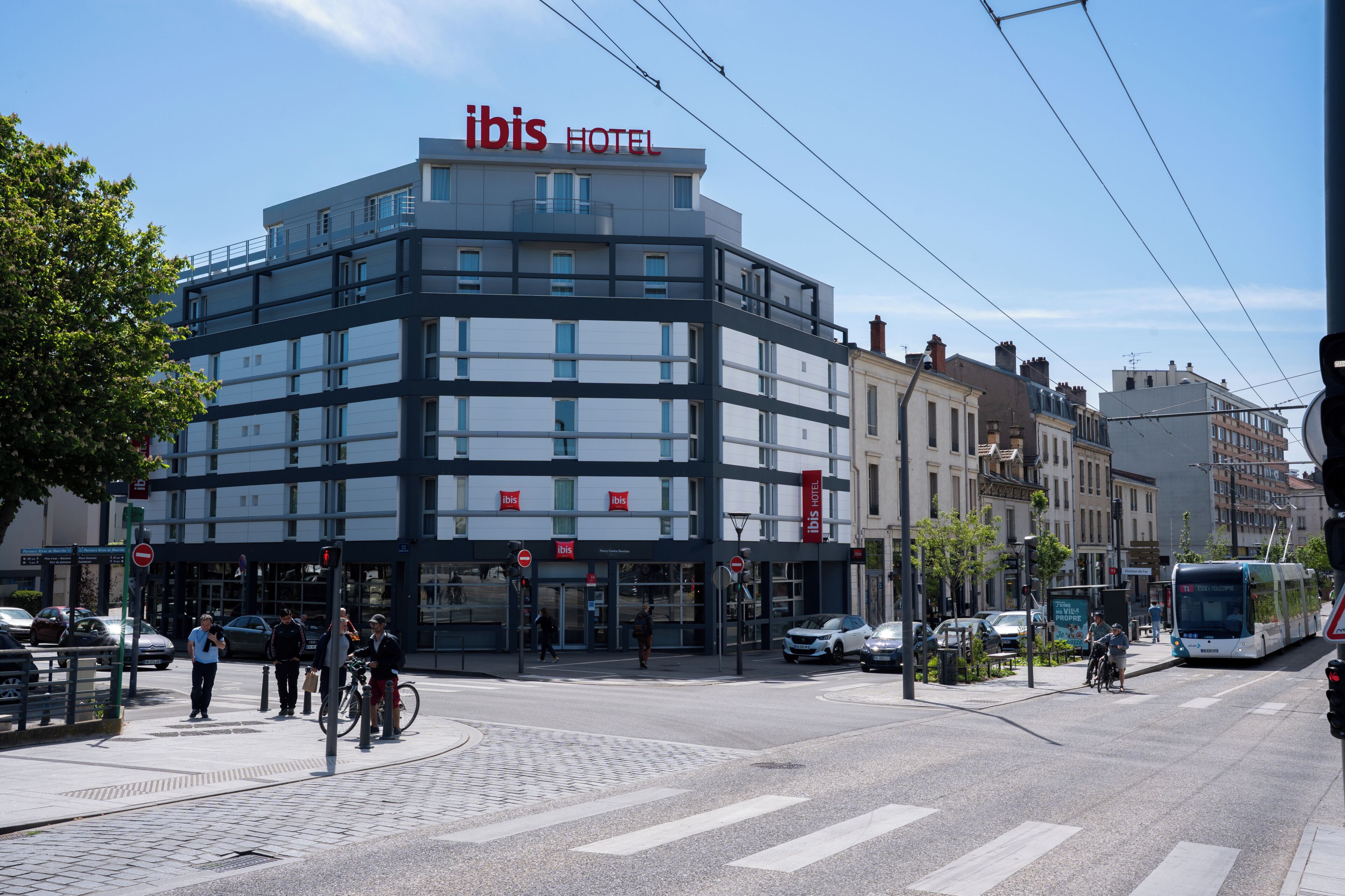 Un hôtel moderne, reconnaissable à son grand panneau « IBIS HOTEL », situé près d’un passage piéton et d’une ligne de tramway.