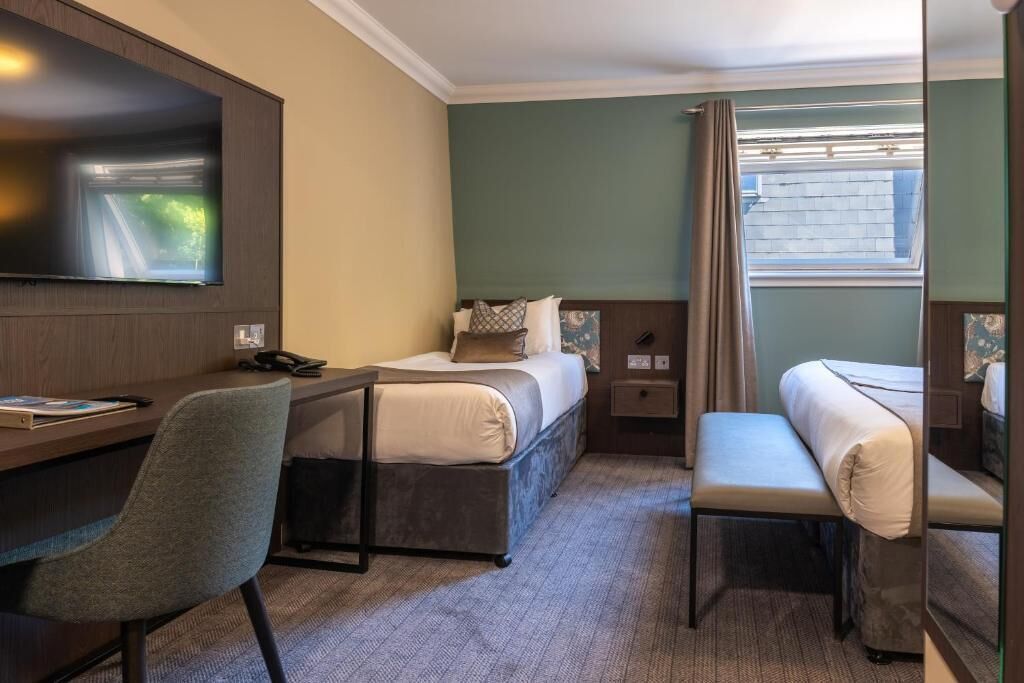 Een hotelkamer met een bed, een bureau met stoel, een aan de muur bevestigde televisie en een raam met gordijnen.