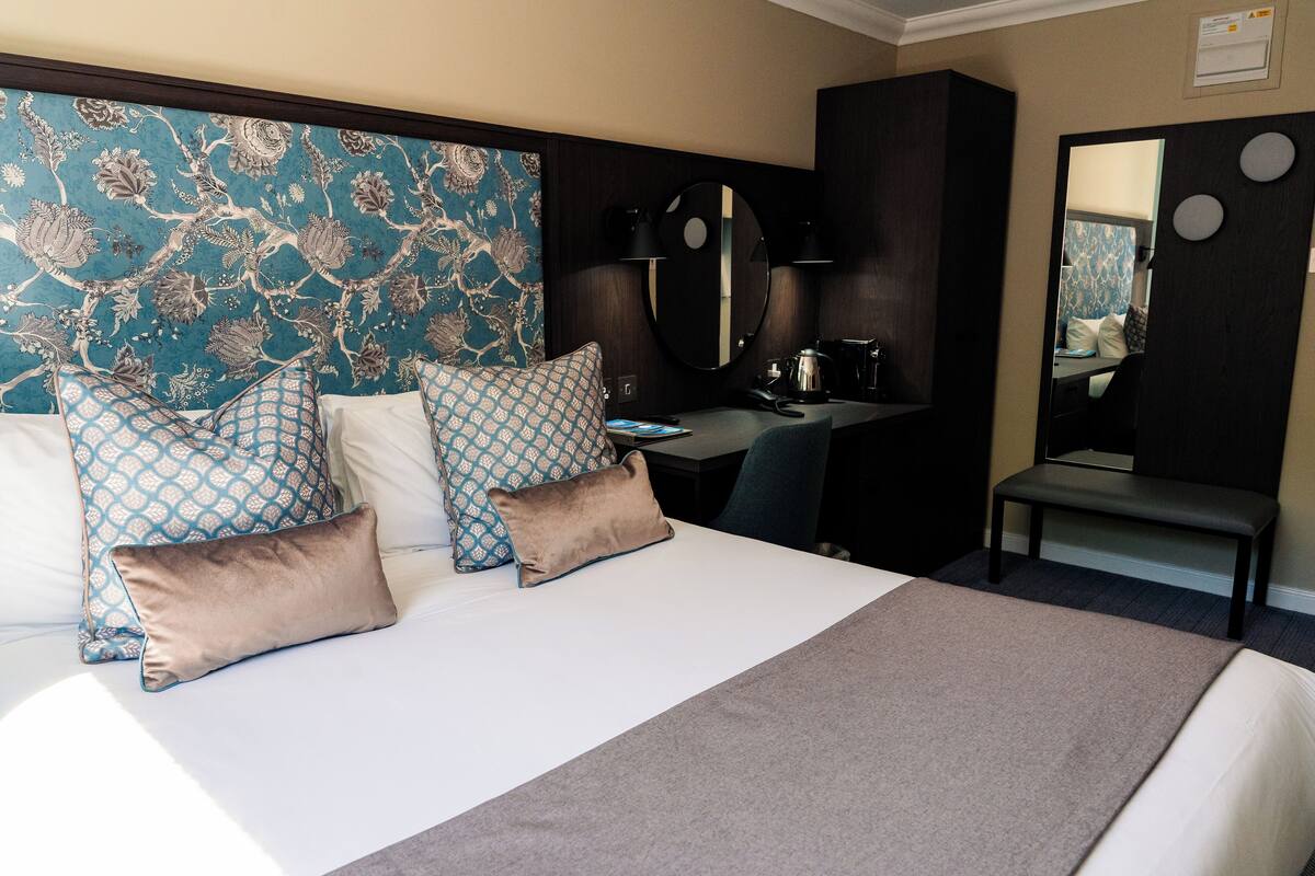 A hotel room with a large bed, two patterned pillows, a bedside table with a coffee maker, a mirror, and a chair.