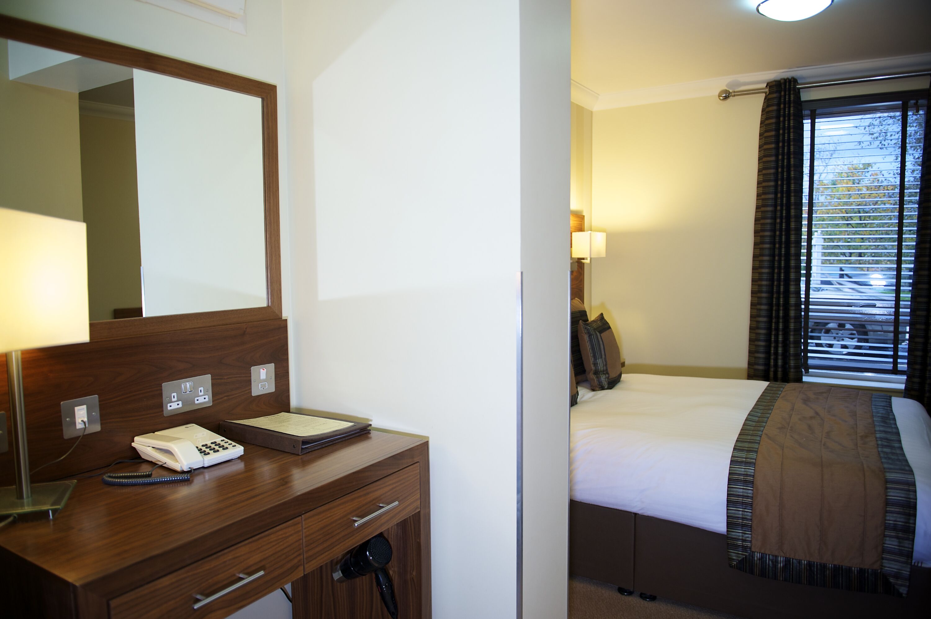 Een hotelkamer met een houten bureau, een telefoon, een lamp en een bed met een bruine plaid.
