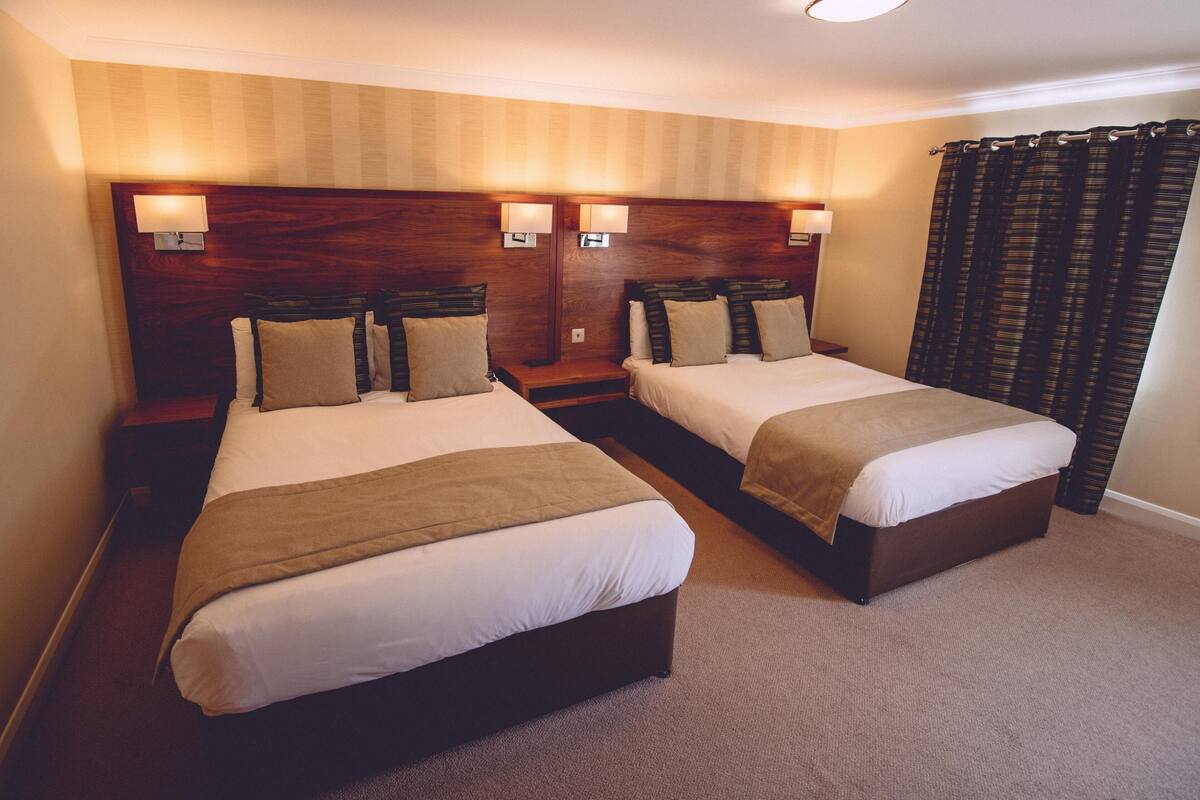 A hotel room with two beds, wooden headboards, bedside lamps, and a carpeted floor.