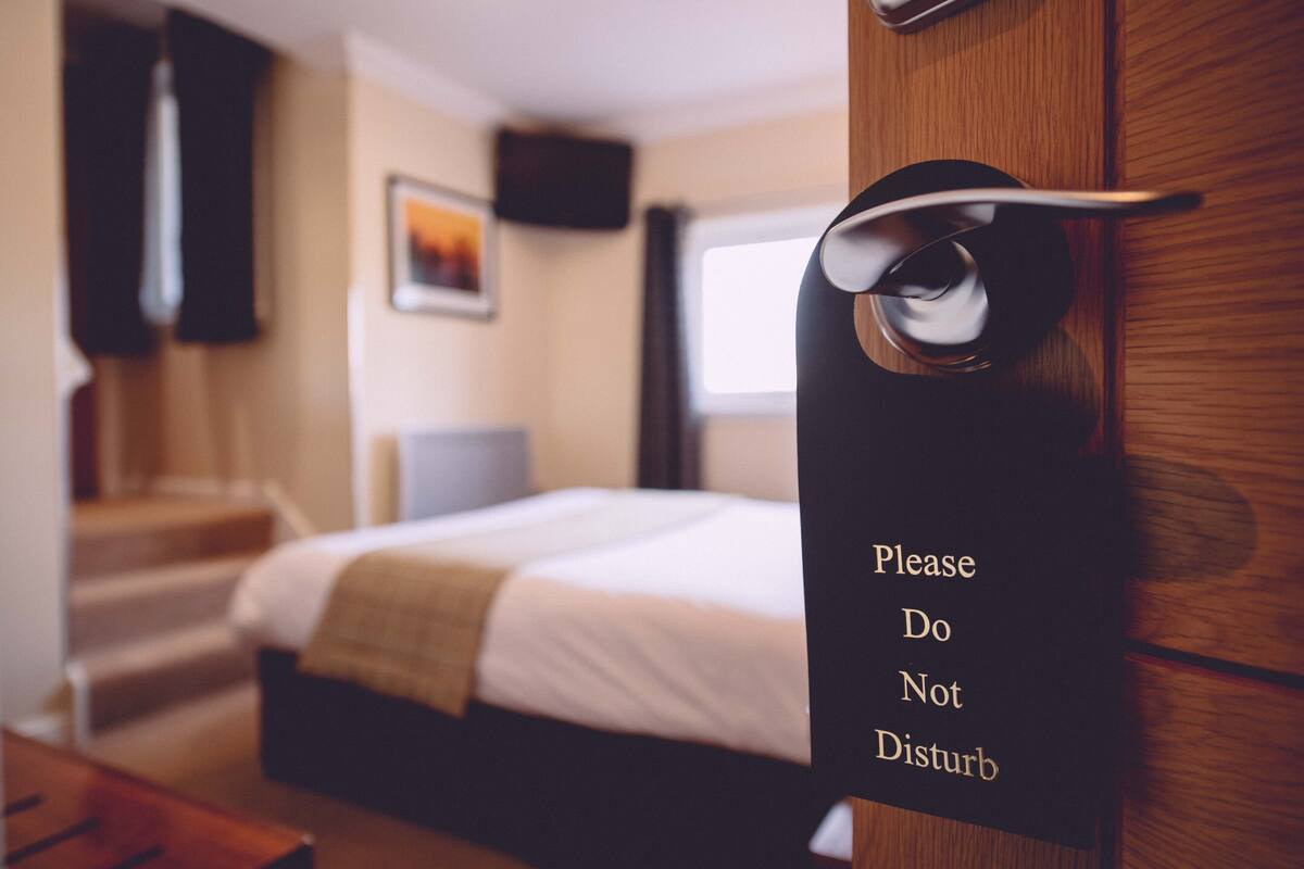 A hotel room with a bed, a bedside table, a TV, and a sign on the door that reads "Please Do Not Disturb".