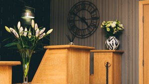 A reception area with a wooden desk, a black leather chair, a vase with flowers, a wall clock, and a door. - Nevis Bank Inn (Fort William)