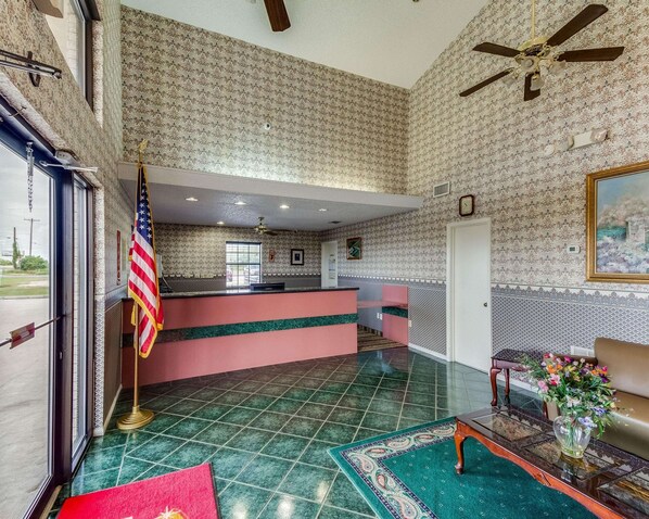 A hotel lobby with a reception desk, a flag, a ceiling fan, and a painting on the wall.