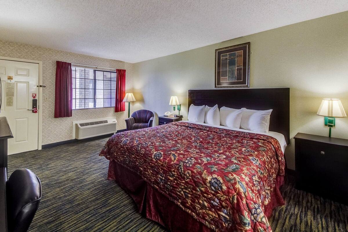 A hotel room with a bed, two bedside lamps, a chair, a desk, a window with curtains, and a framed picture on the wall.