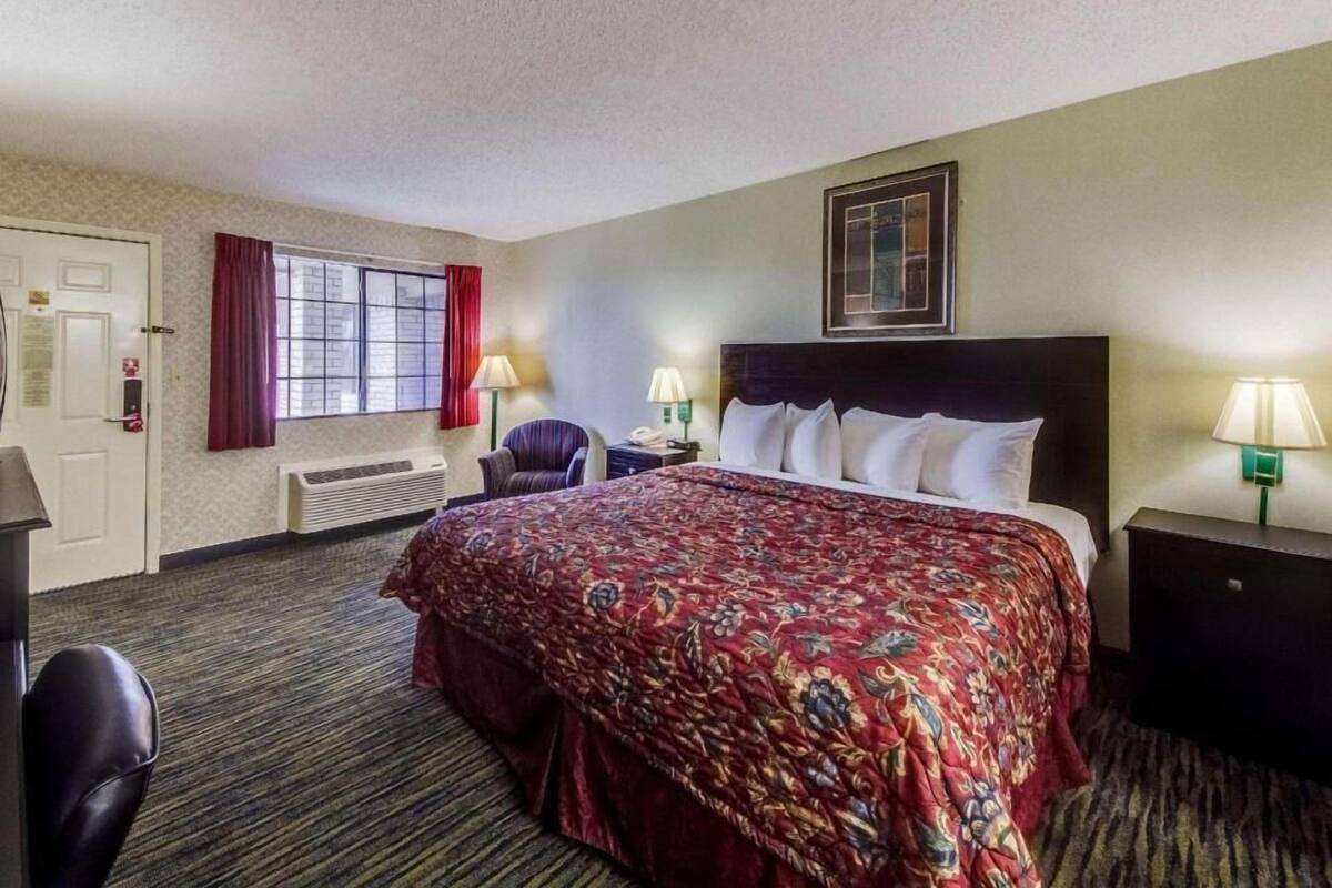 A hotel room with a bed, two bedside lamps, a chair, a desk, a window with curtains, and a framed picture on the wall.