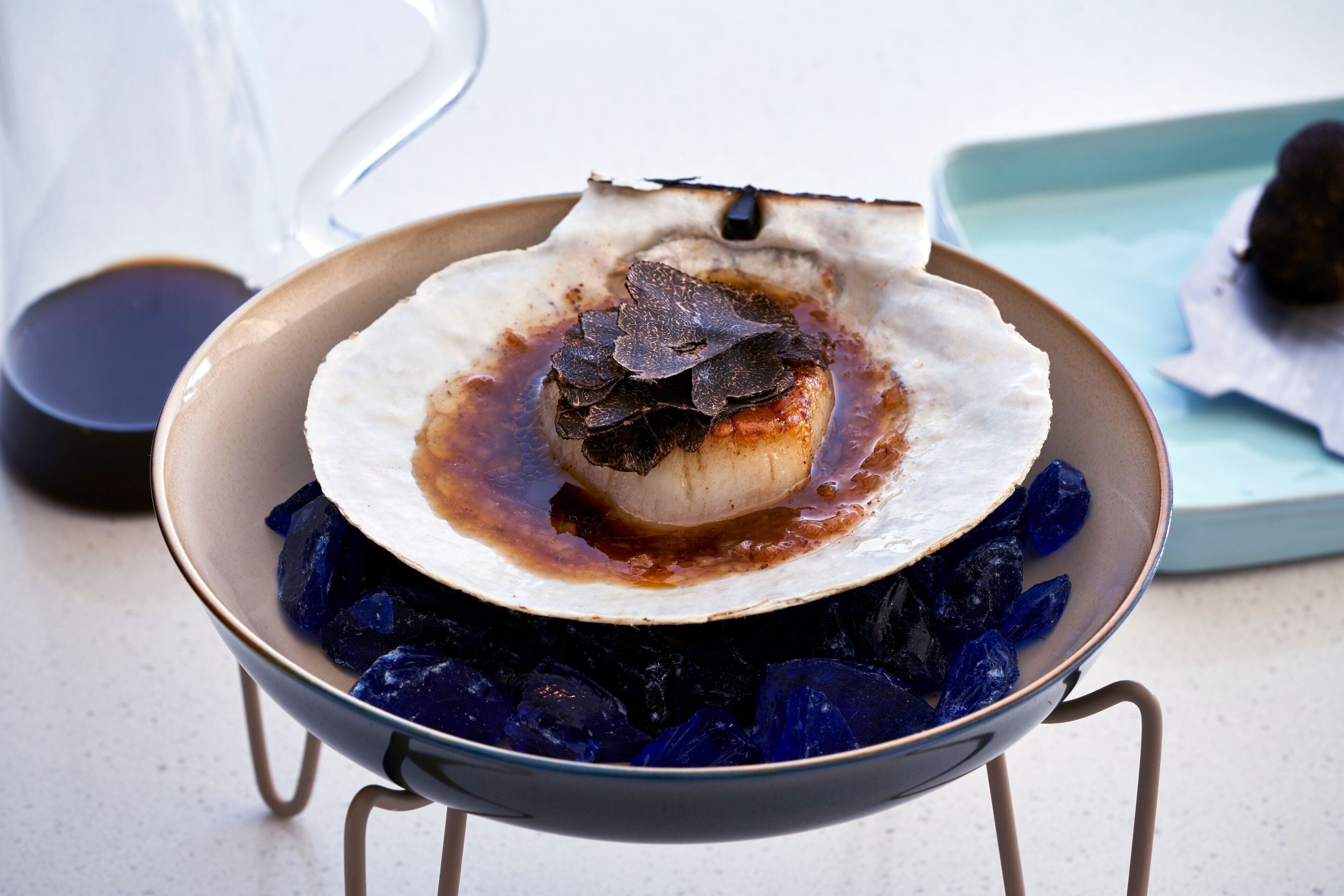 A dish with a white shell, dark leaf garnish, and a sauce, served on a stand with blue crystals.