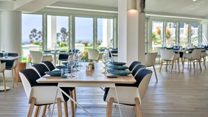 A spacious dining area with a long table set for a meal, surrounded by chairs. Large windows provide a view of the outdoors.