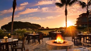 An outdoor dining area with tables and chairs, a central fire pit, and a view of the sunset over a landscape with trees and a building.