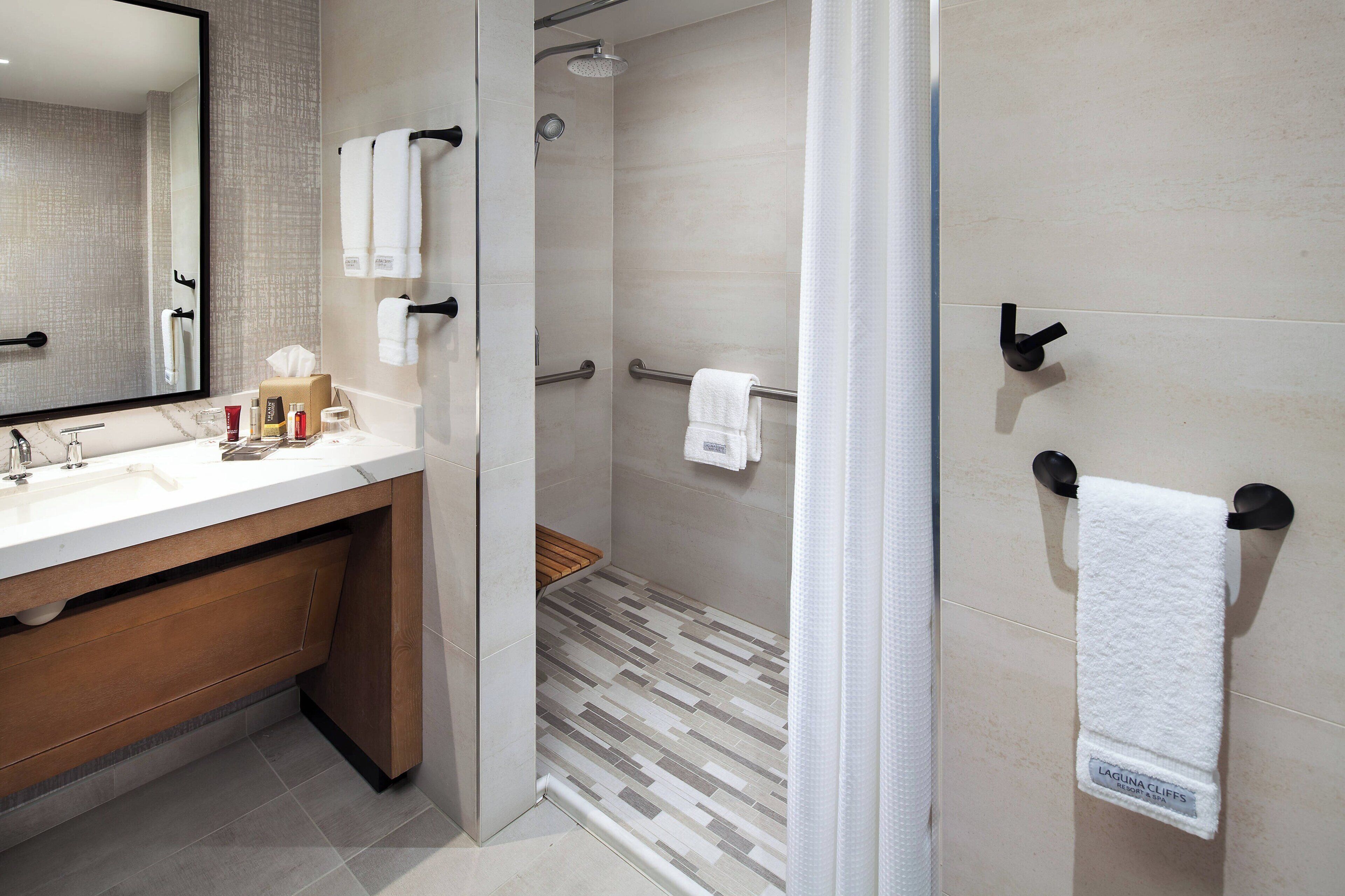 A modern bathroom with a large mirror, a sink with toiletries, a shower with a glass door, and a towel rack with a white towel.