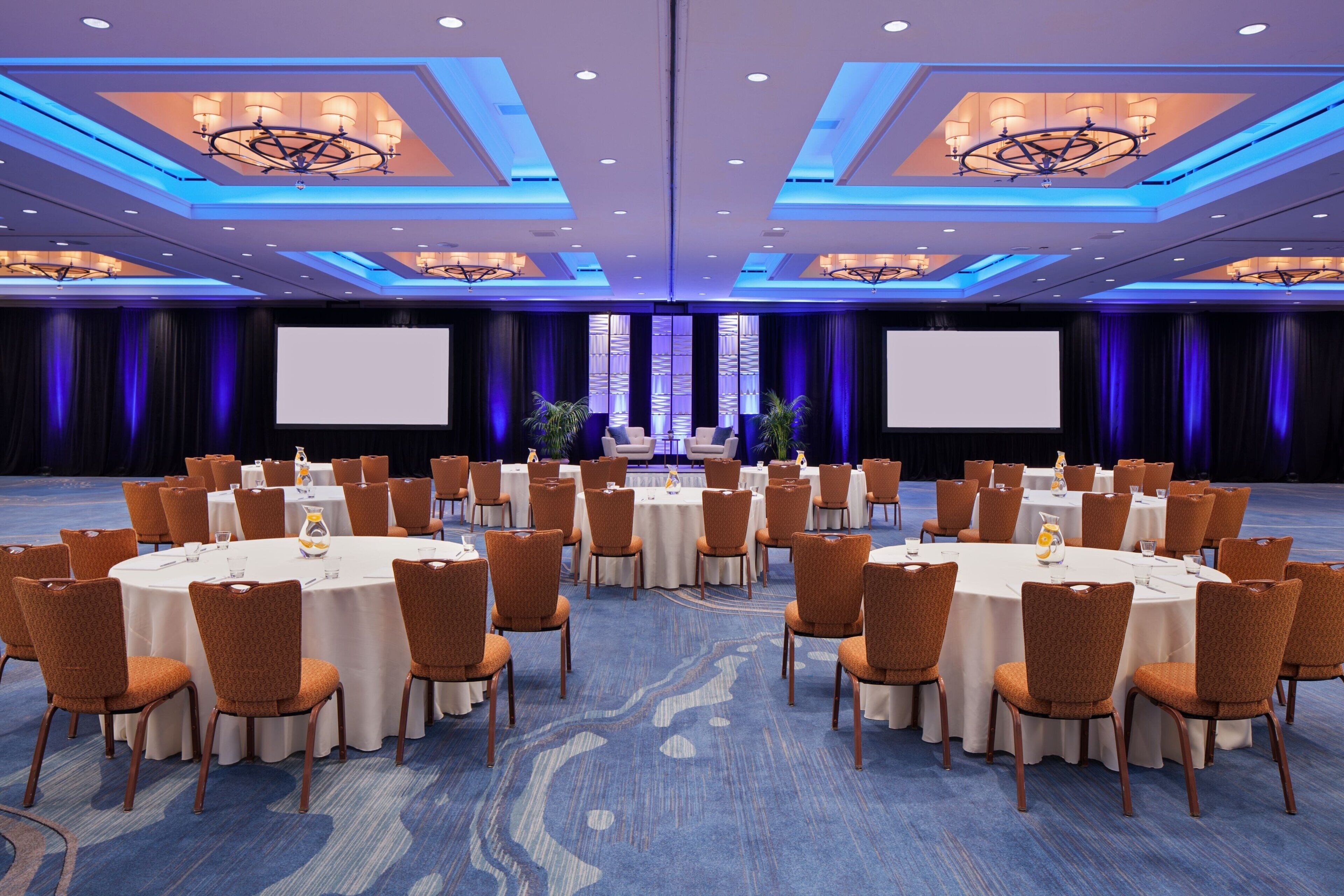 A large conference room with round tables and chairs, a stage with a large screen, and a blue carpeted floor.