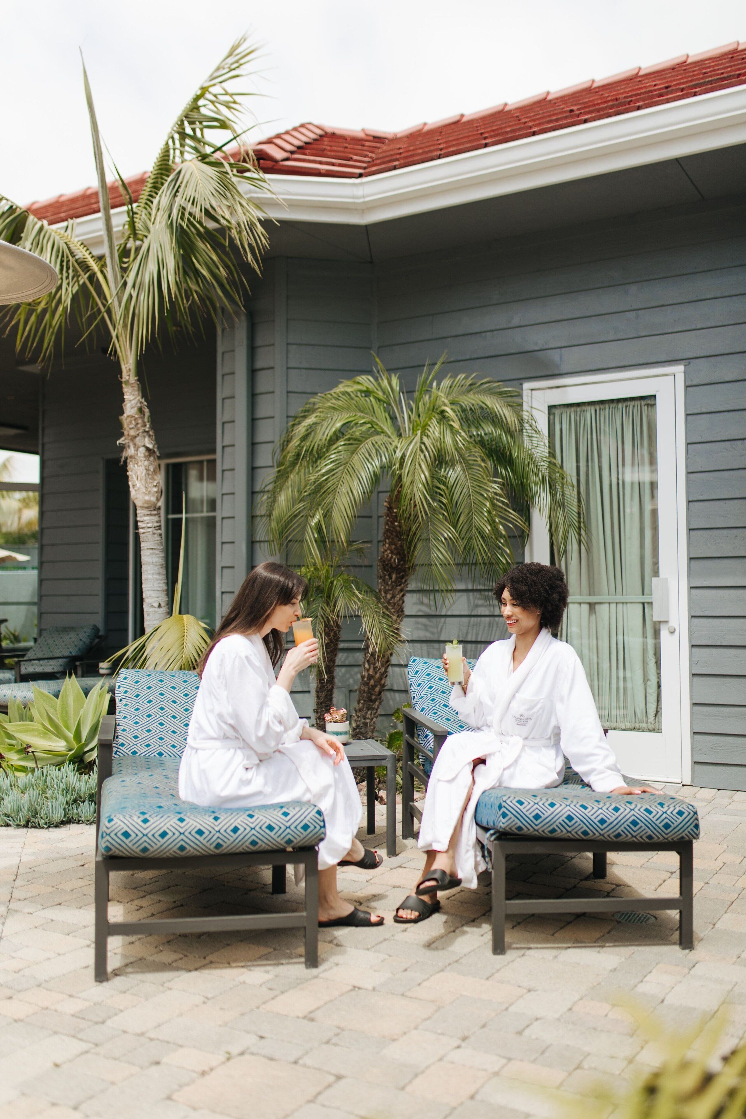 Two people sitting on patterned chairs outside a building with a red roof, palm trees, and a window with curtains.