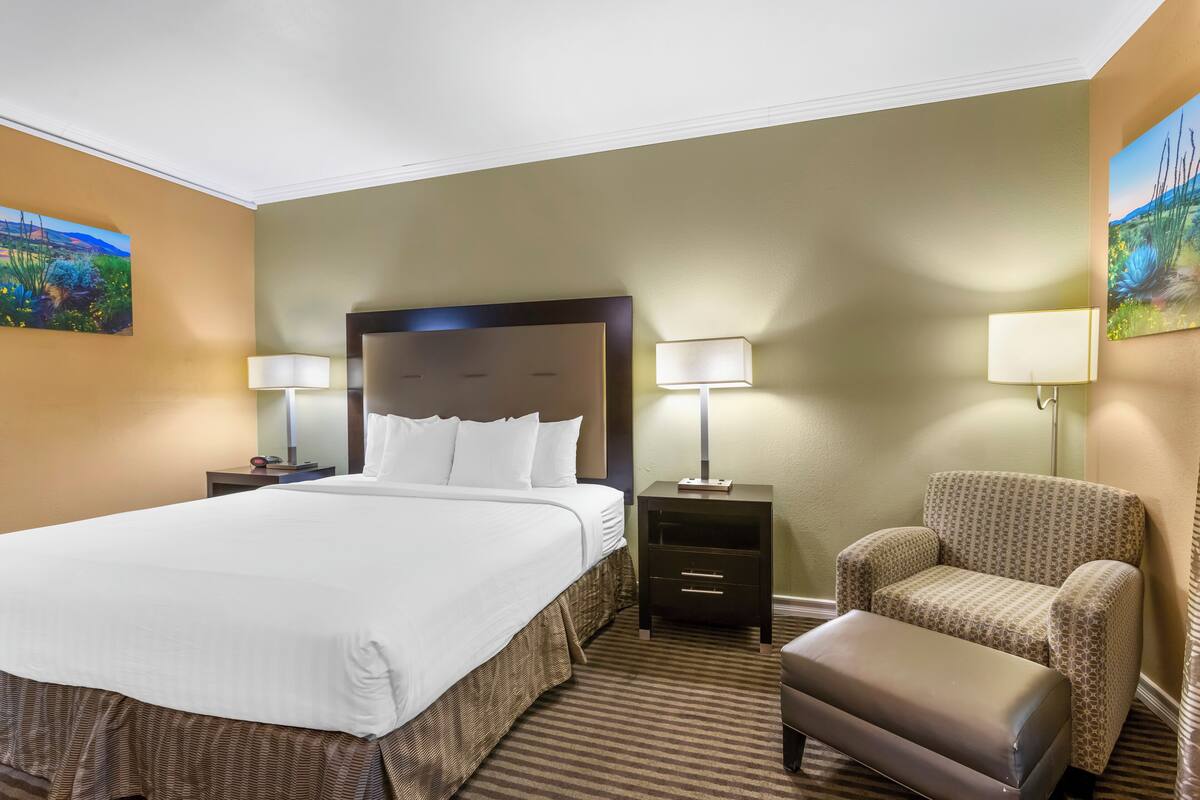 A hotel room with a large bed, two bedside tables with lamps, a patterned armchair, and a small ottoman.