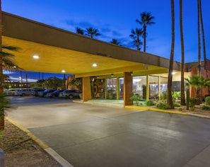 A hotel entrance with a covered parking area, palm trees, and a clear sky. - Royal Sun Hotel & Suites (Tucson)