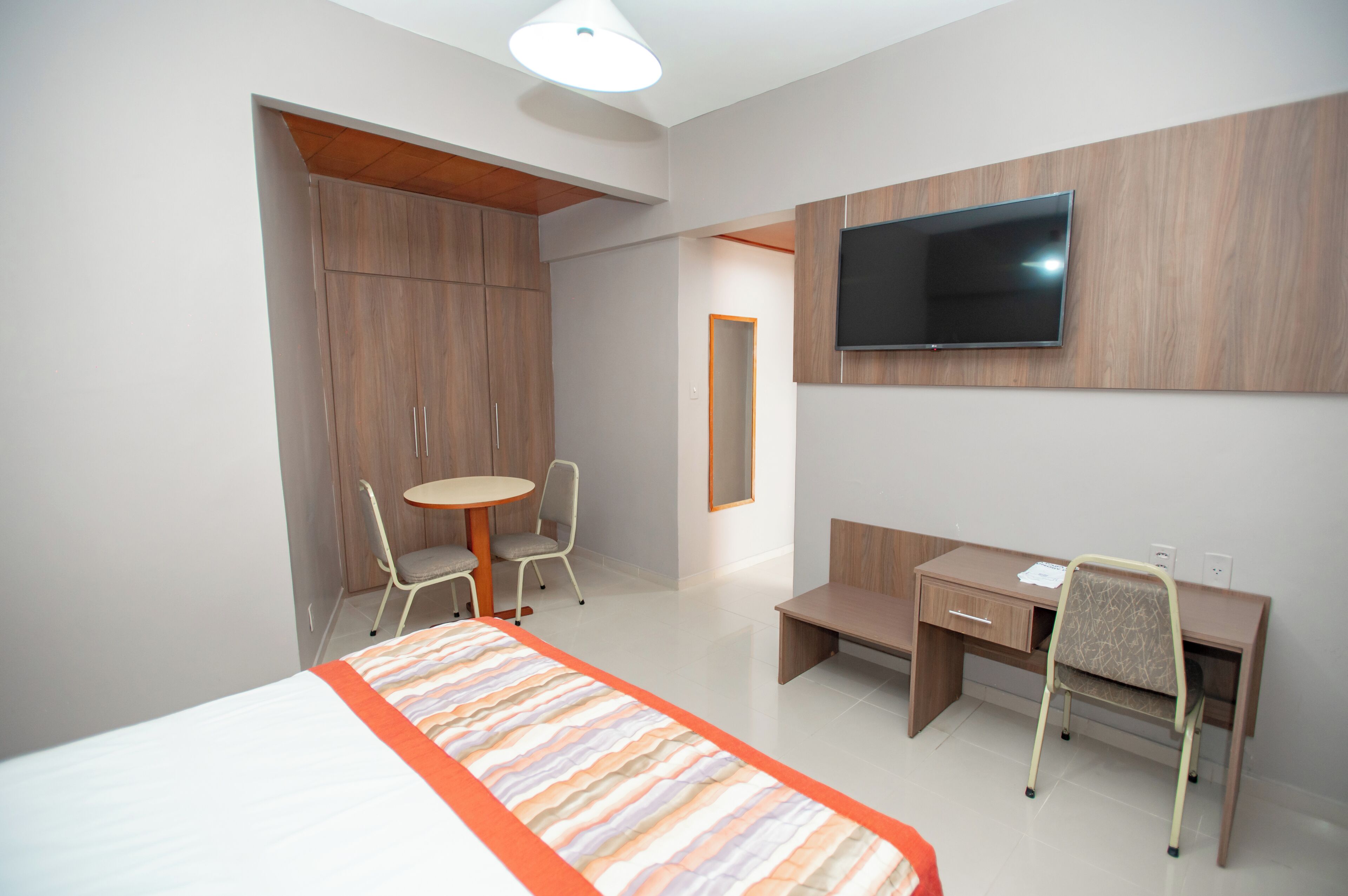 A hotel room with a bed, a desk, a chair, a small table, and a TV mounted on the wall.