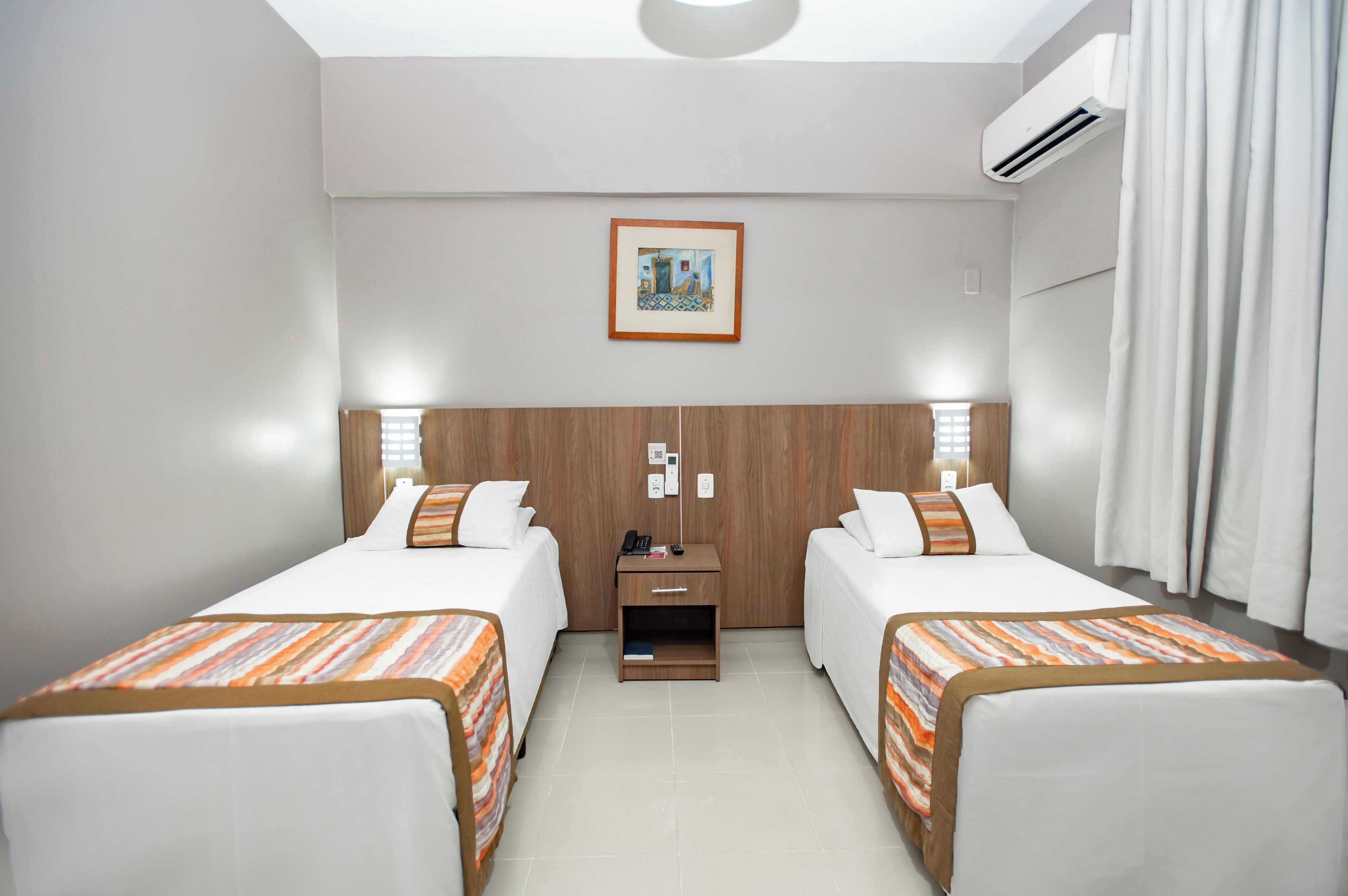 A hotel room with two beds, a wooden headboard, a nightstand with a phone and books, and an air conditioning unit.