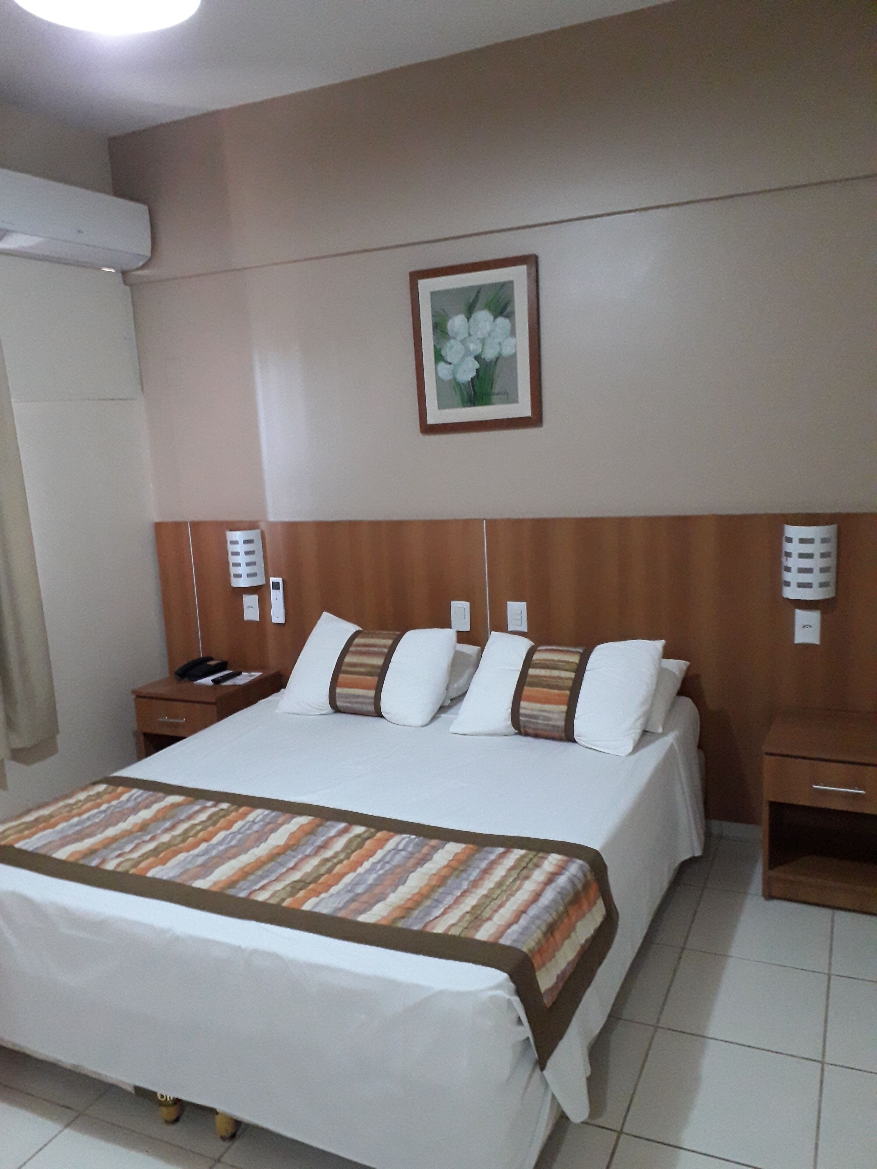 Quarto de hotel com uma cama grande, cabeceira de madeira, mesas de cabeceira, telefone e uma moldura com um quadro na parede.