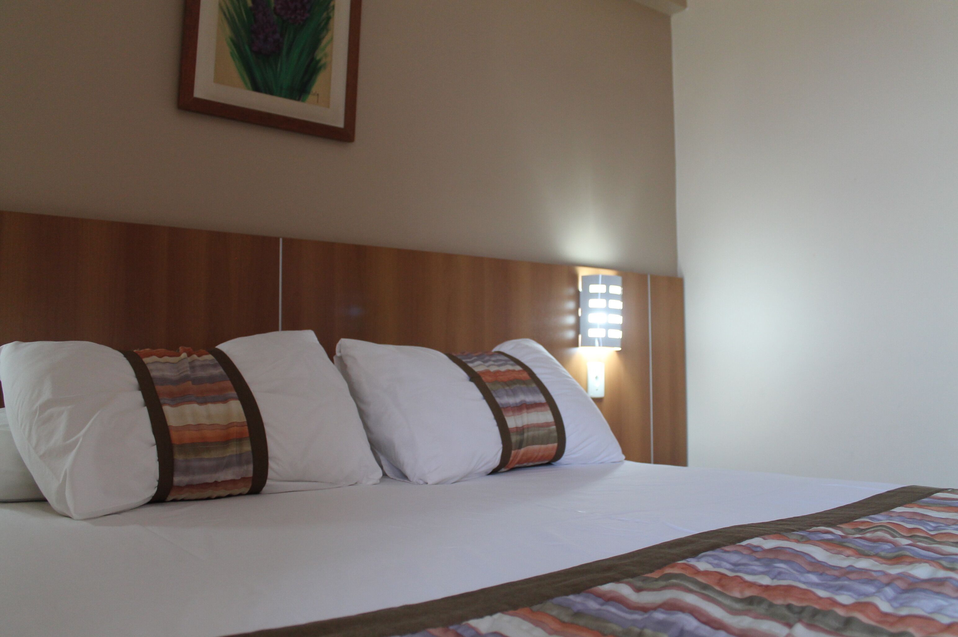 A neatly made bed with white linens and striped pillows, a wooden headboard, and a framed picture on the wall.