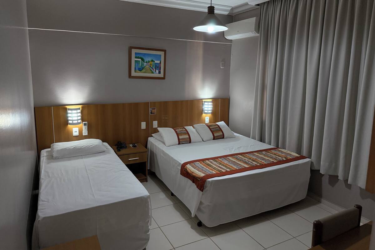 A hotel room with two single beds, a large bed, a painting on the wall, and a window with curtains.