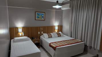 Habitación de hotel con dos camas individuales, una cama grande, un cuadro en la pared y una ventana con cortinas.