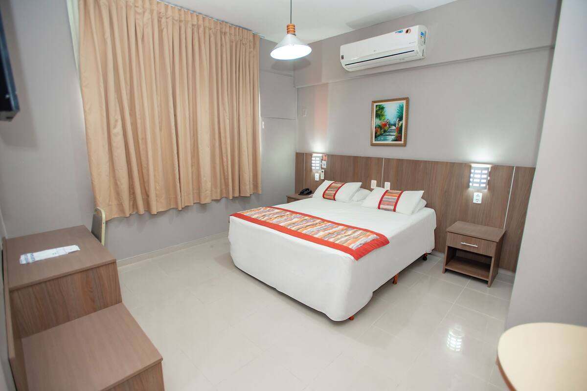 A hotel room with a bed, bedside tables, a desk, a chair, an air conditioner, and a window with curtains.