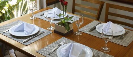A dining table set with white plates, folded napkins, and wine glasses, with a potted plant and red flowers in the center.