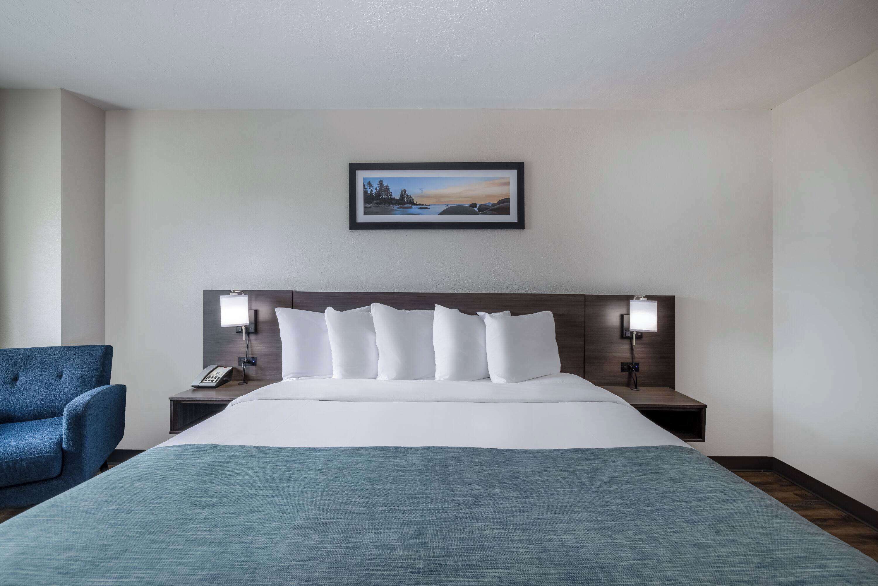 A hotel room with a large bed, a blue armchair, a nightstand with a lamp, and a framed picture on the wall.
