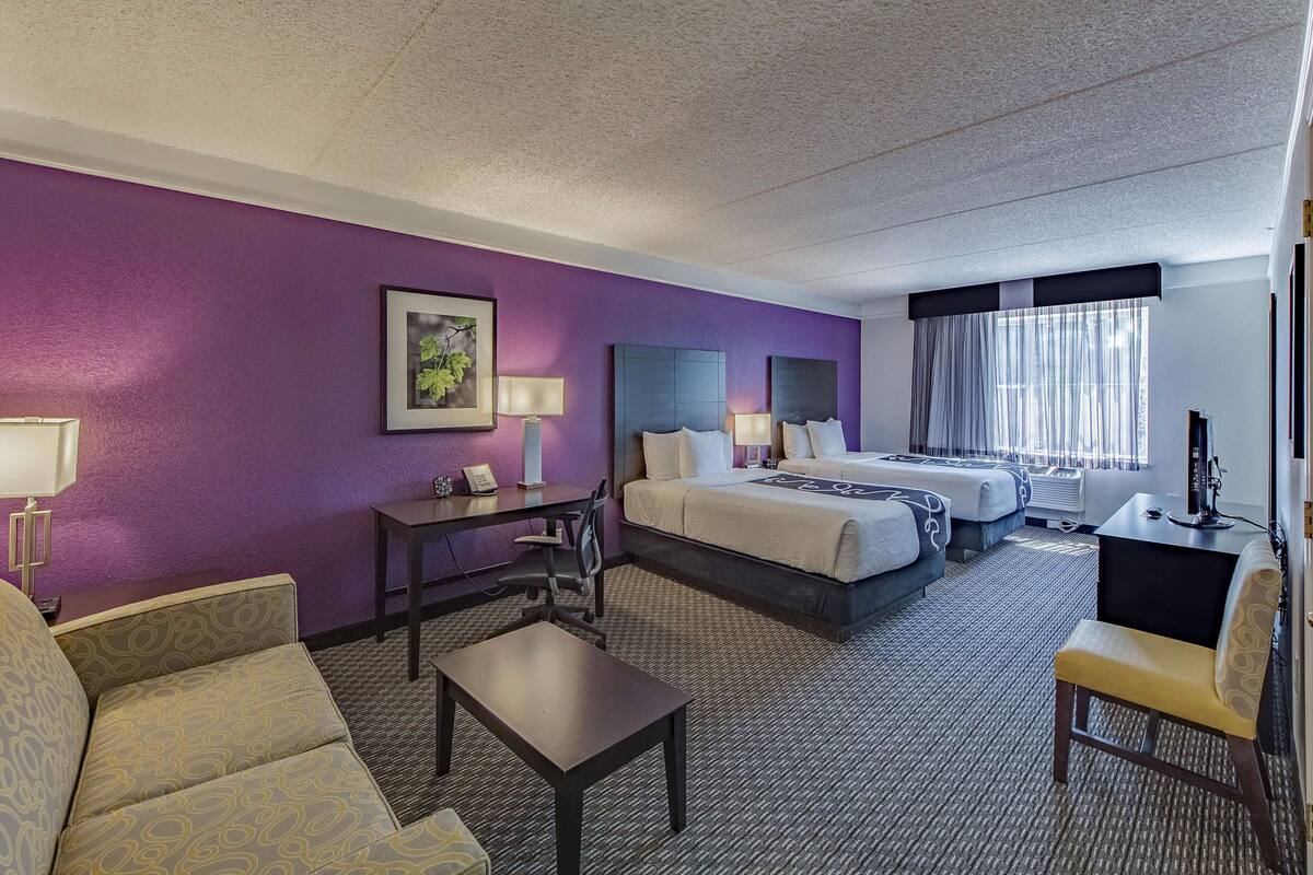 Una habitación de hotel con una pared morada, una cama, un escritorio, una silla y un sofá.