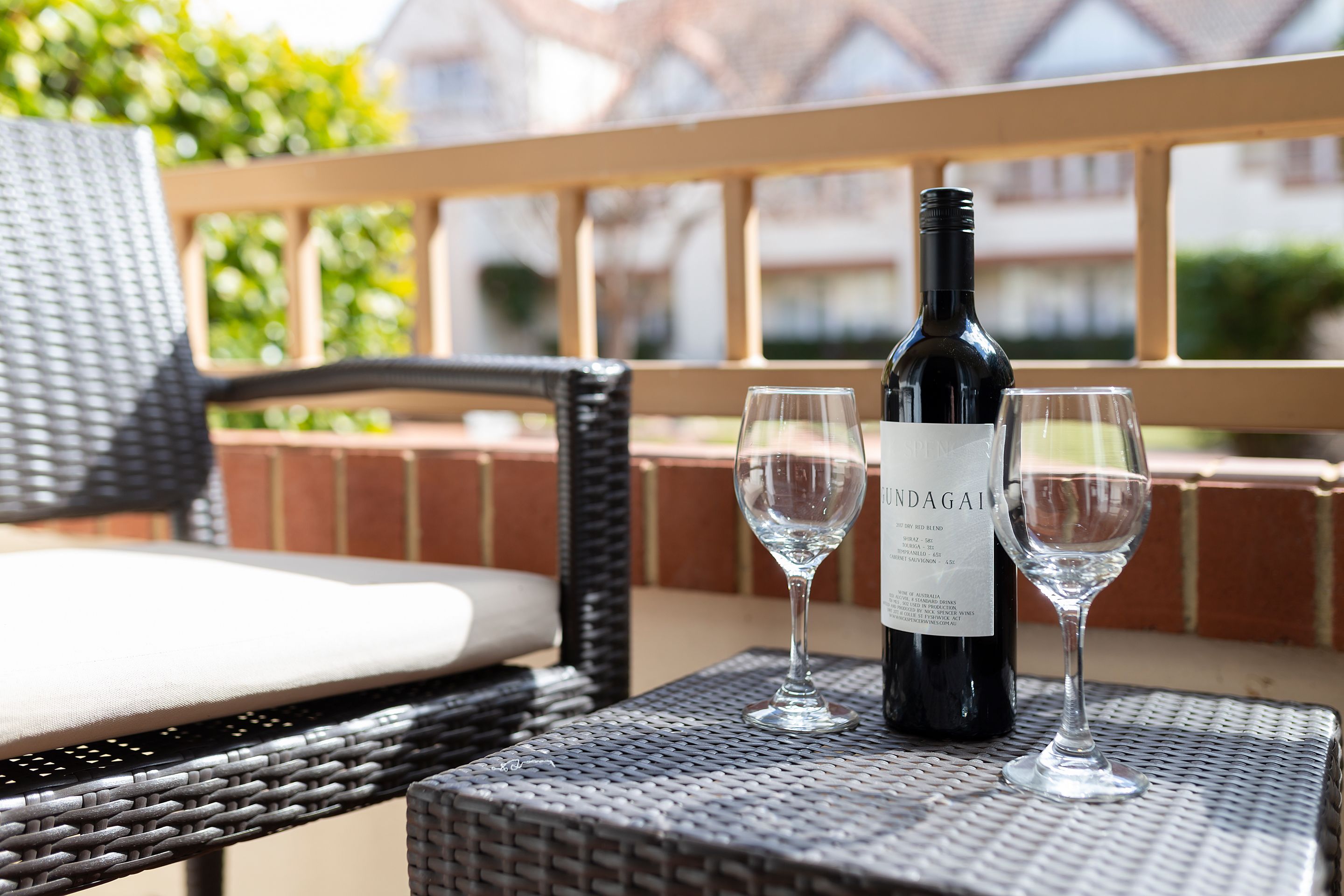 A wicker chair with a cushion, a bottle of wine, and two glasses on a table.