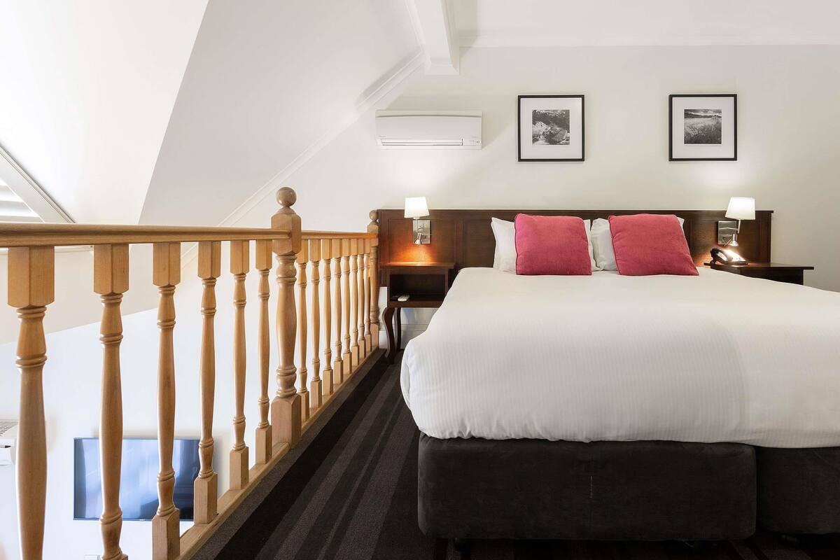 A hotel room with a large bed, two bedside tables with lamps, a wooden staircase railing, and two framed pictures on the wall.
