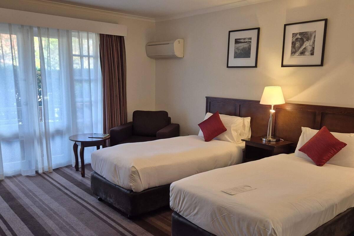 A hotel room with two beds, a chair, a small table, and a lamp.