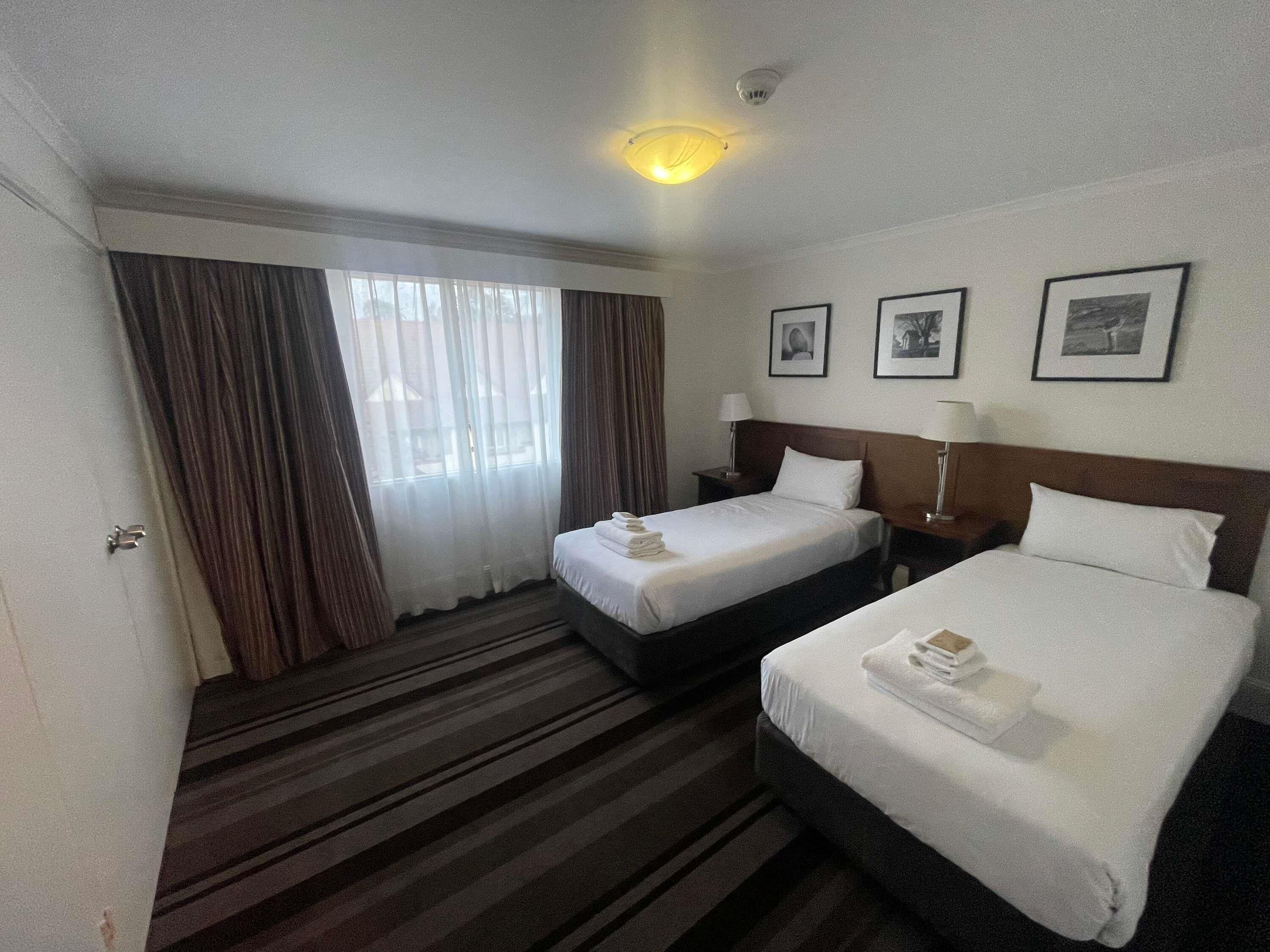 Une chambre d’hôtel avec deux lits, une grande fenêtre avec des rideaux, un luminaire au plafond et trois tableaux encadrés accrochés au mur.