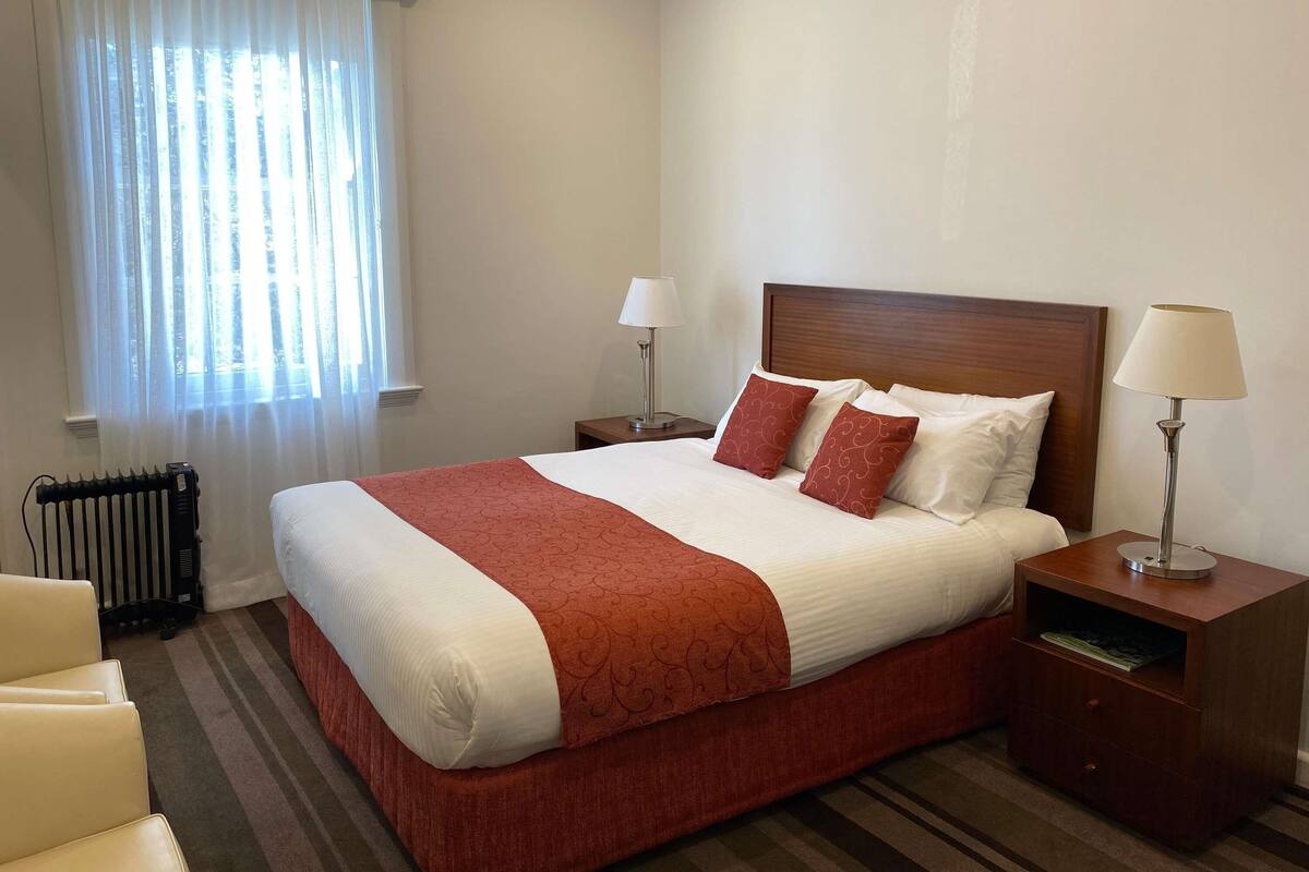 A hotel room with a large bed, two bedside tables, a radiator, and a window with curtains.