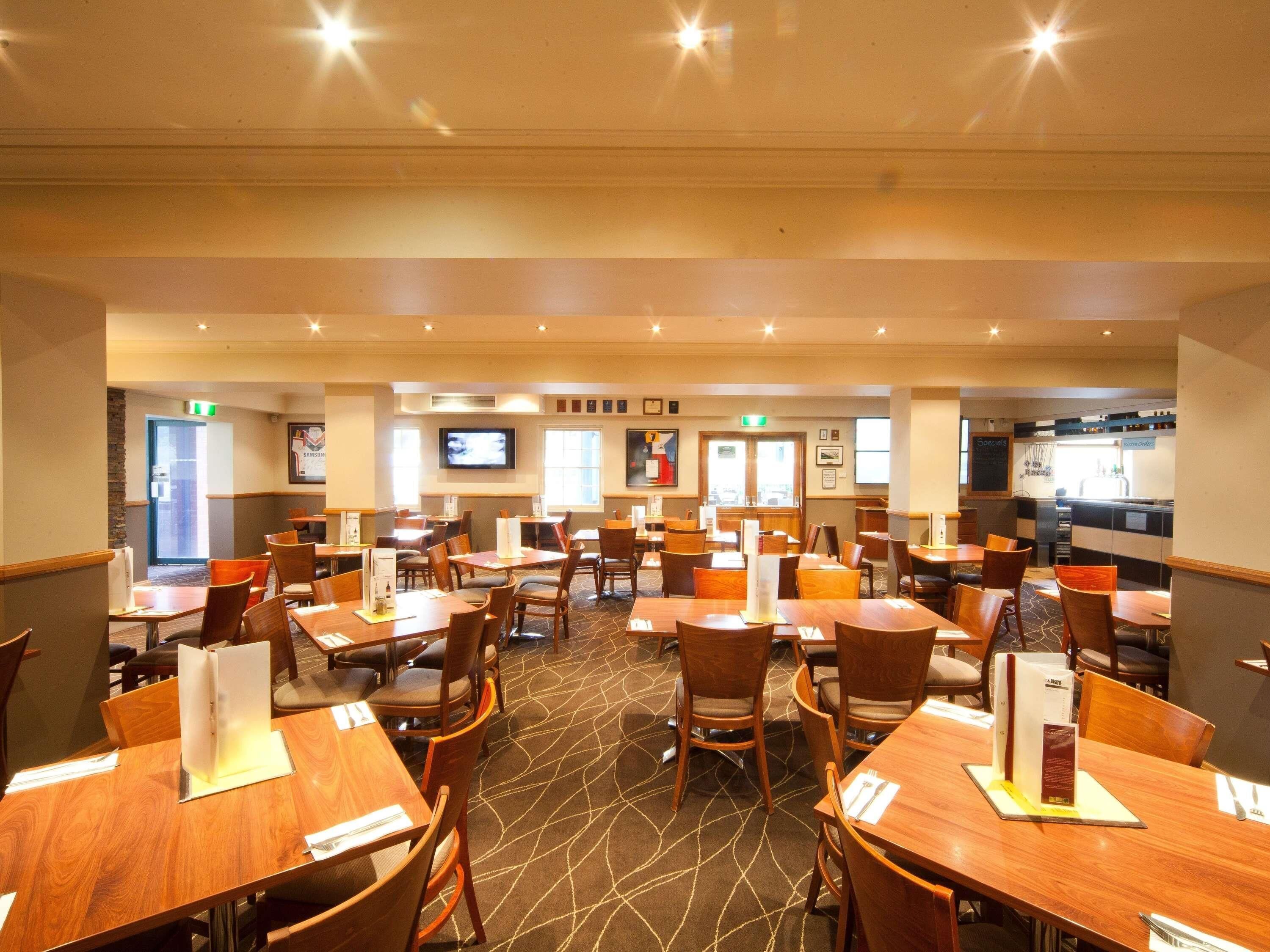 A hotel dining area with multiple tables and chairs, a television on the wall, and a kitchen area in the background.