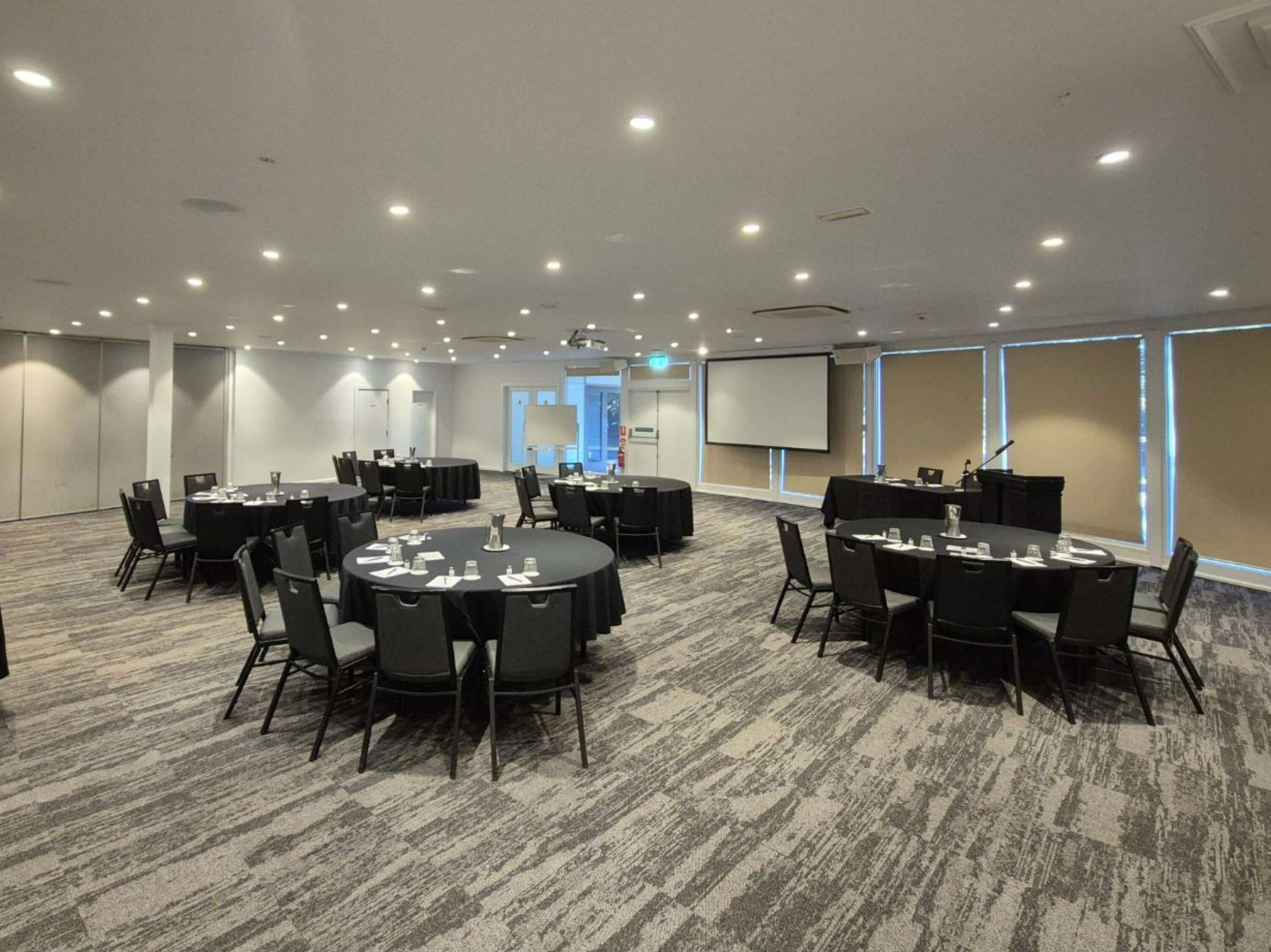 A conference room with multiple tables and chairs, a projector screen, and a carpeted floor.
