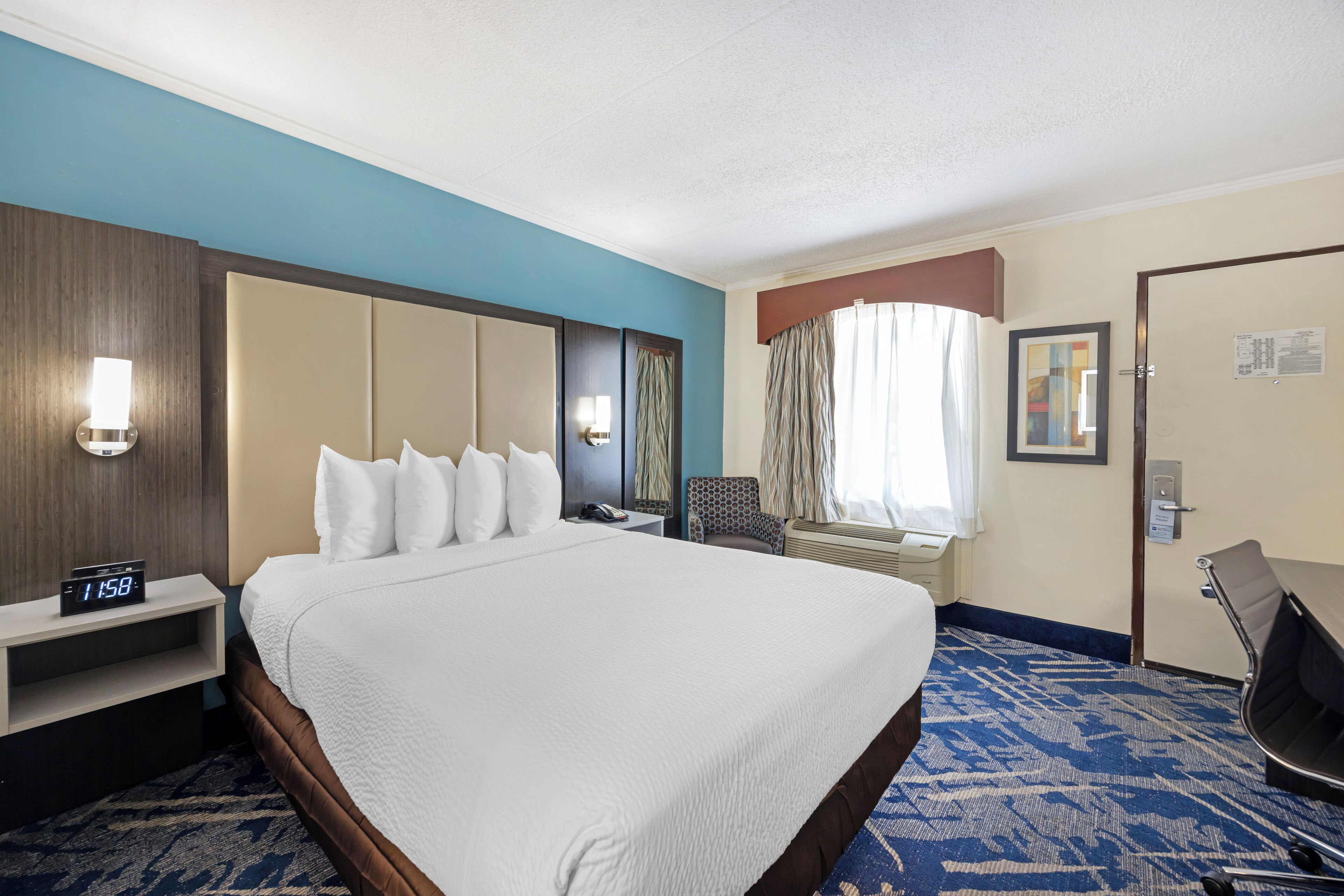 A hotel room with a large bed, a bedside table with a clock, a chair, a desk, and a window with curtains.