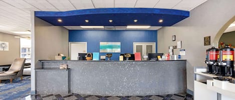 A hotel reception area with a blue accent wall, a reception desk, a coffee station, and a seating area with a patterned floor.