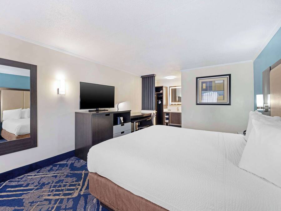 A hotel room with a large bed, a flat-screen TV, a desk with a chair, a mirror, and a bathroom visible through an open door.