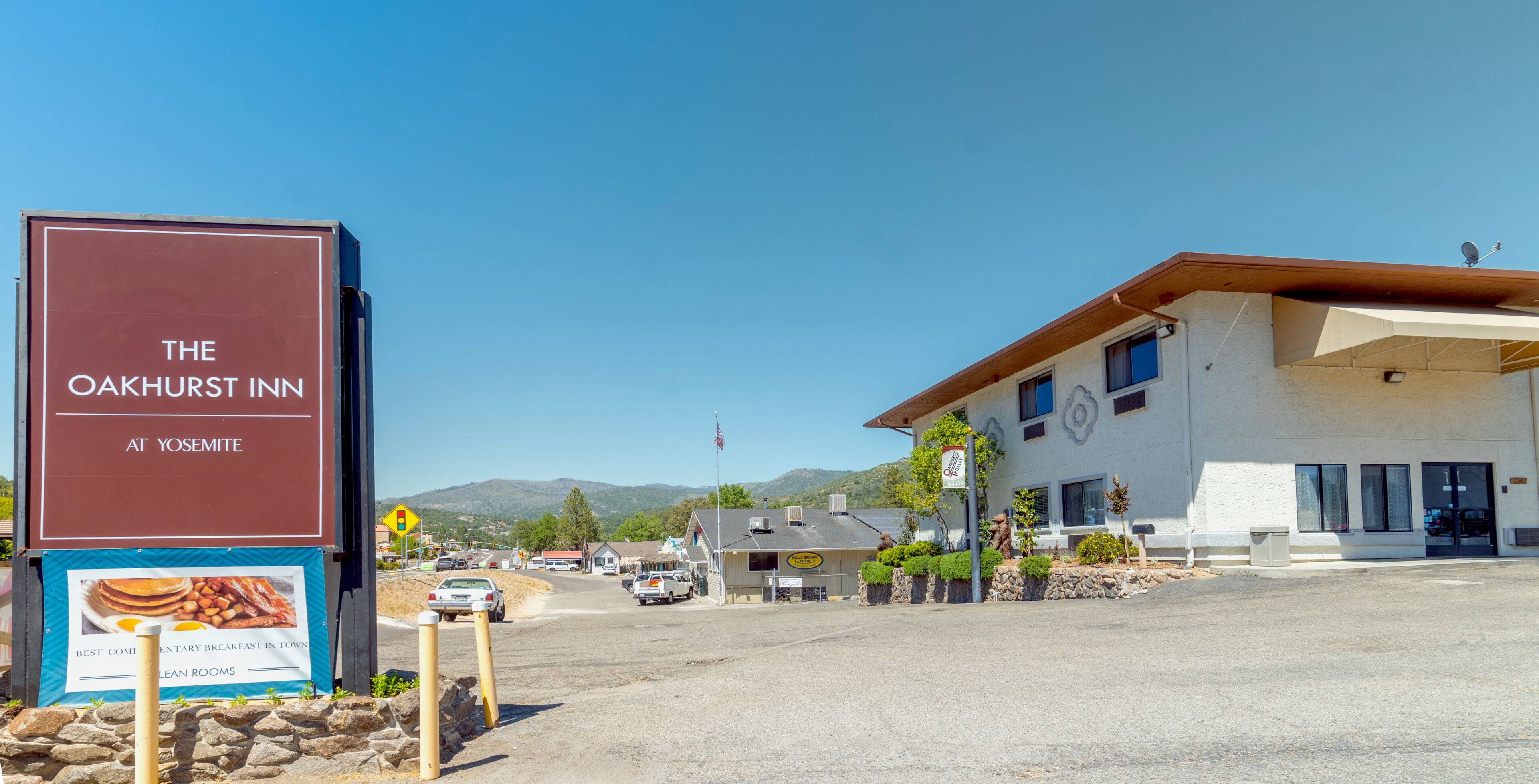 A building with a sign for The Oakhurst Inn at Yosemite, a paved road, and a mountainous background.