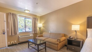 Ein Hotelzimmer mit einer Couch, einem Glastisch, einem Nachttisch mit Lampe, einem Heizkörper und einem Fenster mit Vorhängen.