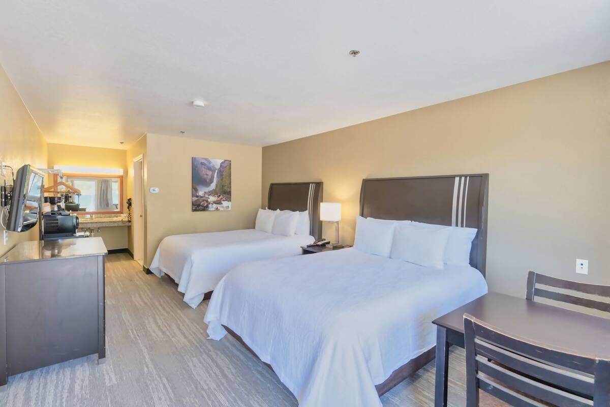 A hotel room with two beds, a desk, a chair, a television, and a bathroom visible in the background.