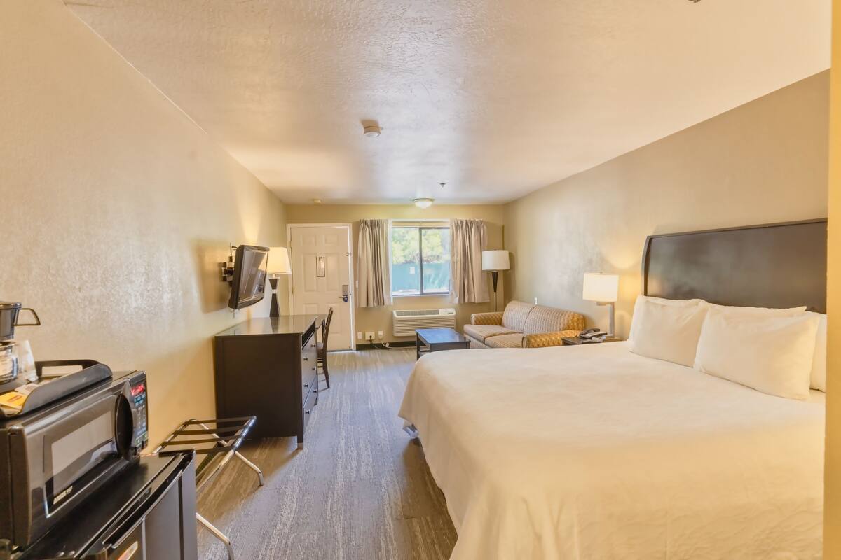 A hotel room with a bed, desk, chair, TV, and a small table with a lamp.