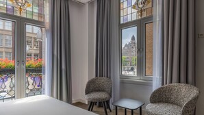 Ein Hotelzimmer mit einem Bett, zwei gemusterten Sesseln, einem kleinen Tisch und großen Fenstern mit Vorhängen.