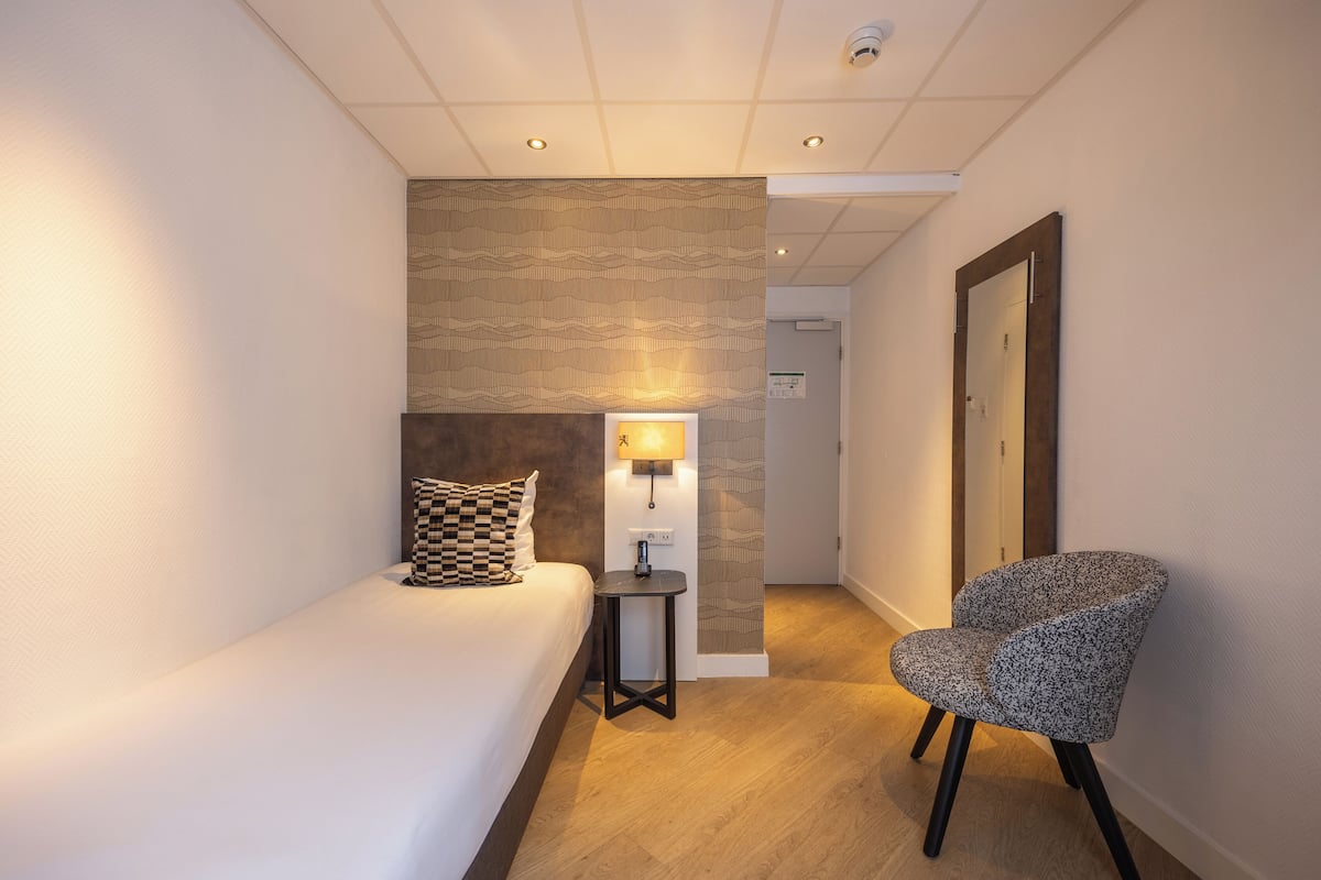 A hotel room with a bed, a chair, a small table with a lamp, and a patterned wall.