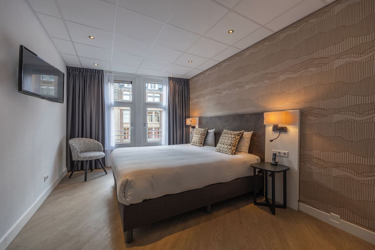 A modern hotel room with a large bed, a chair, a bedside table with a lamp, and a patterned wall.