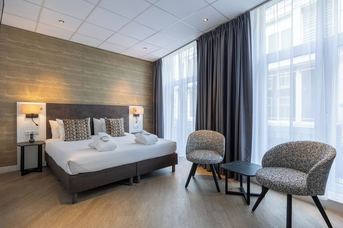 A modern hotel room with a large bed, two bedside tables with lamps, a small table, two patterned armchairs, and a large window with curtains.