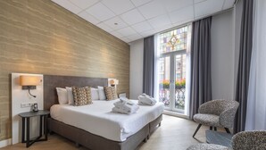 Ein modernes Hotelzimmer mit einem großen Bett, einem kleinen Beistelltisch, einem Sessel und Blick ins Freie durch ein großes Fenster.