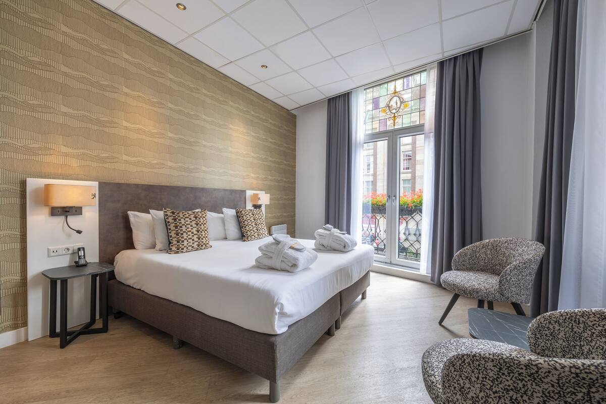 A modern hotel room with a large bed, a small side table, a chair, and a view of the outside through a large window.